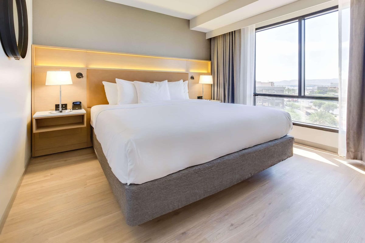Habitación de hotel con una cama grande, lámparas de noche, una mesita de noche y vistas de la ciudad a través de un ventanal amplio.