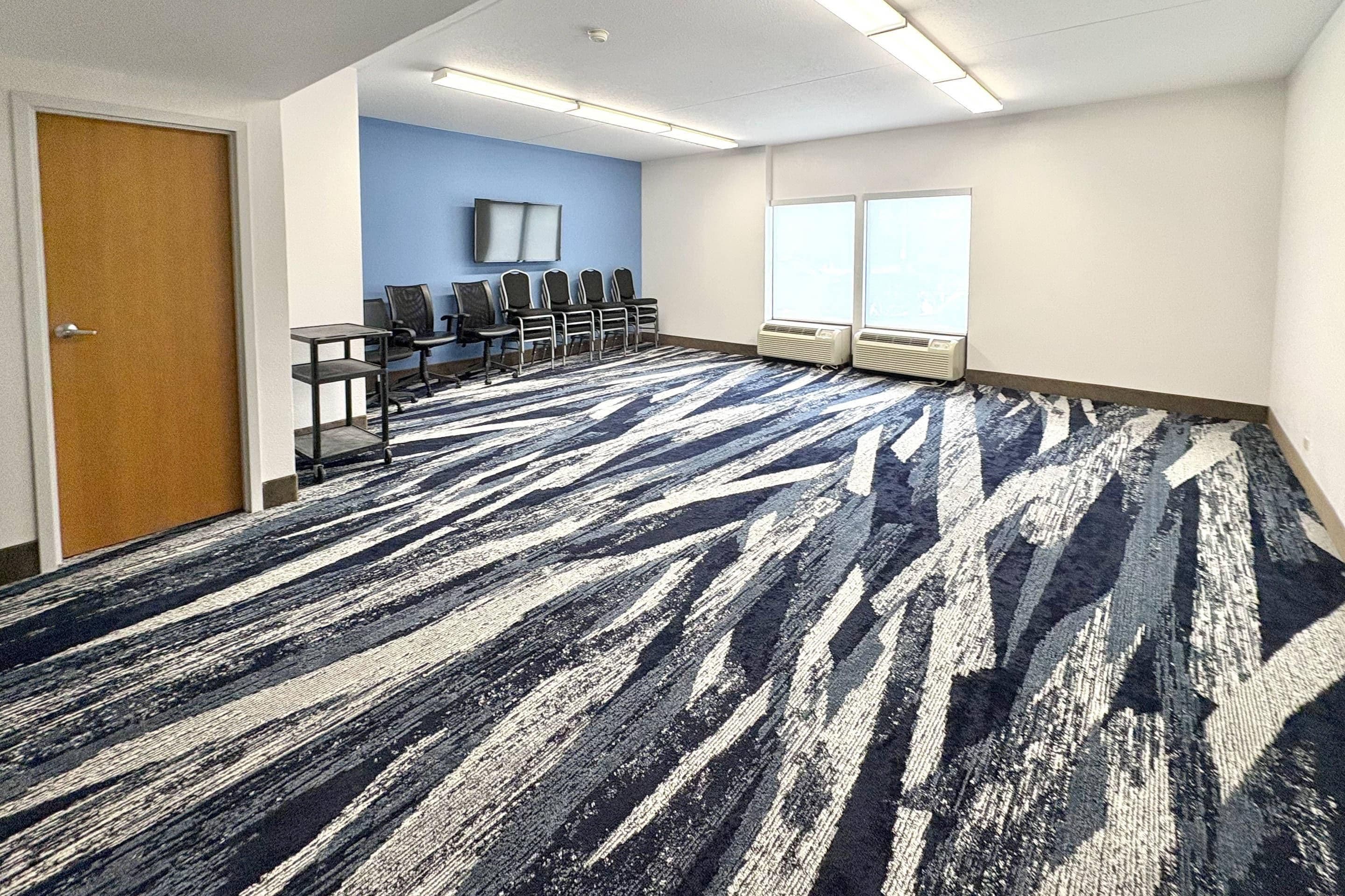 A conference room with a patterned carpet, a row of chairs, a television on the wall, and large windows.