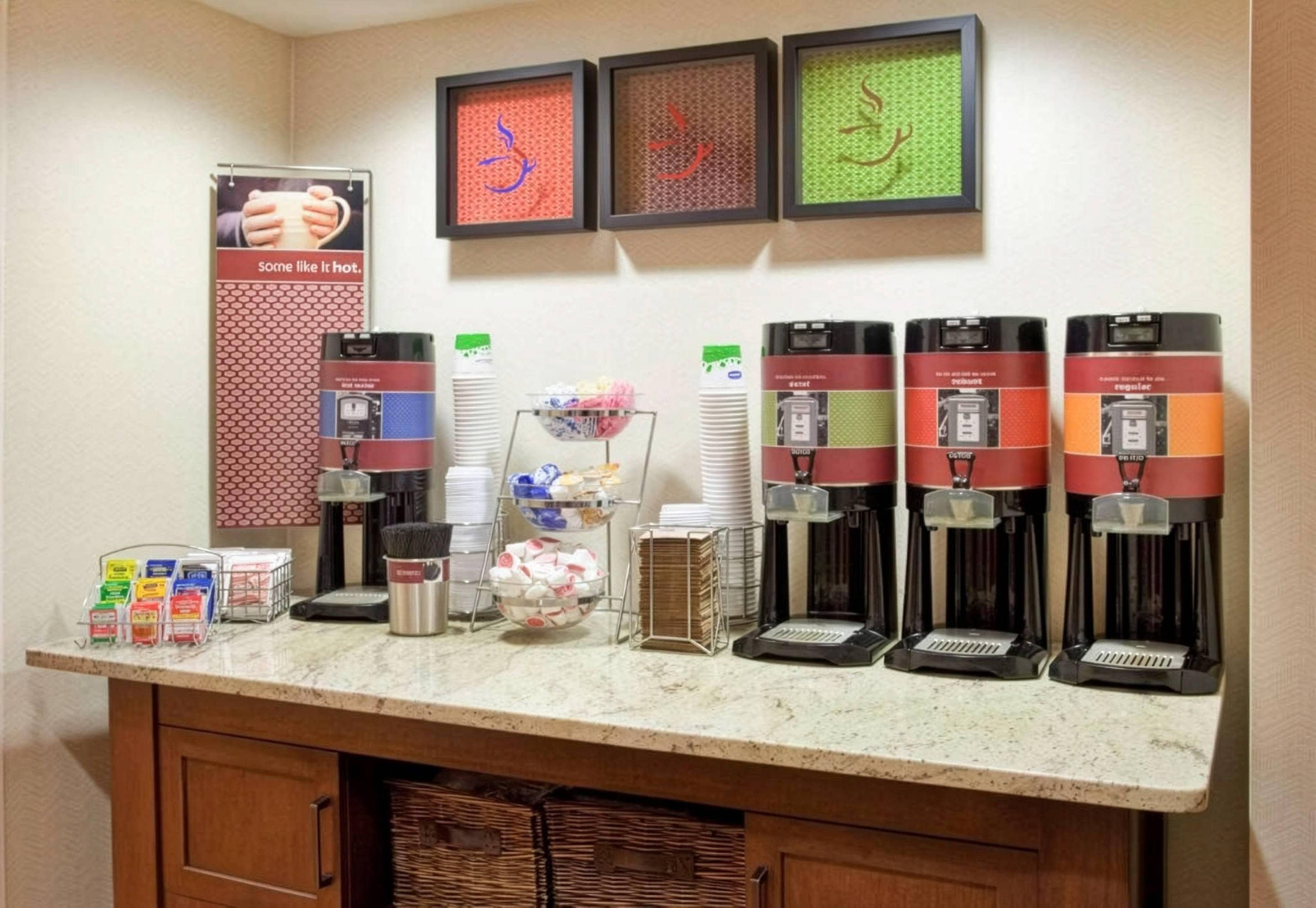 Uma estação de café com várias máquinas de café, um letreiro indicando bebidas quentes e uma parede com obras de arte emolduradas.