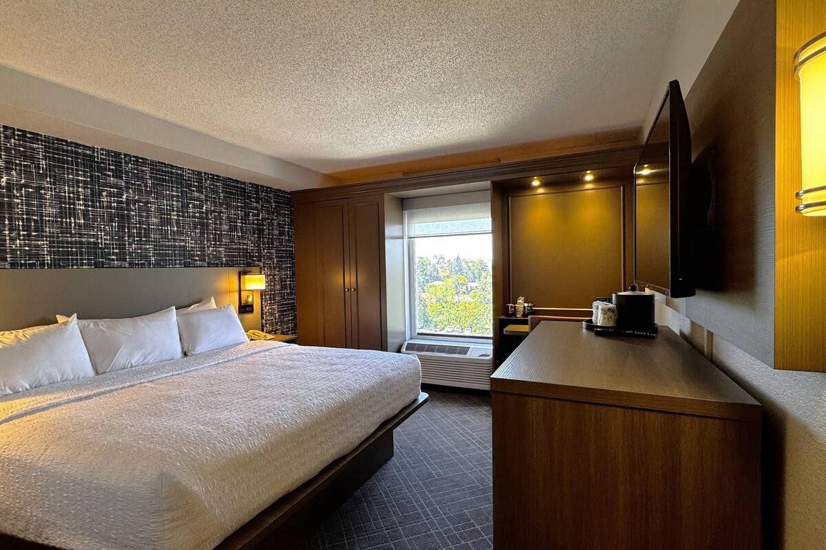 A hotel room with a large bed, a desk, a mirror, and a window with a view of trees.