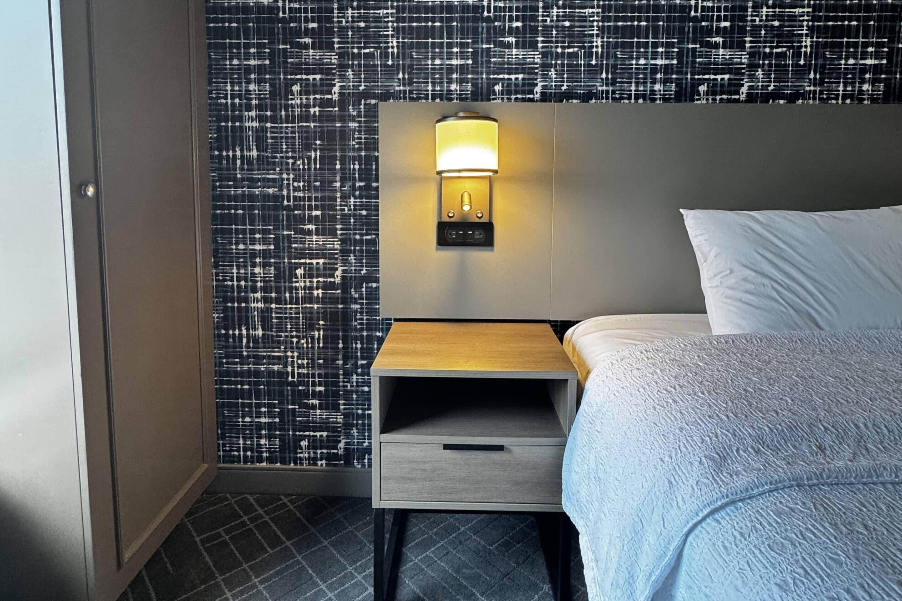 A hotel room with a bed, a nightstand with a lamp, a small drawer, and a patterned wall.
