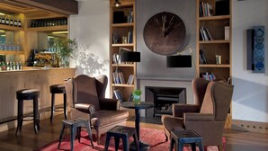 Un salón acogedor con sillones de cuero, una mesa de centro y una alfombra roja. Se observa una estantería con libros y un reloj grande en la pared. En el fondo, hay una zona de bar con botellas y vasos.