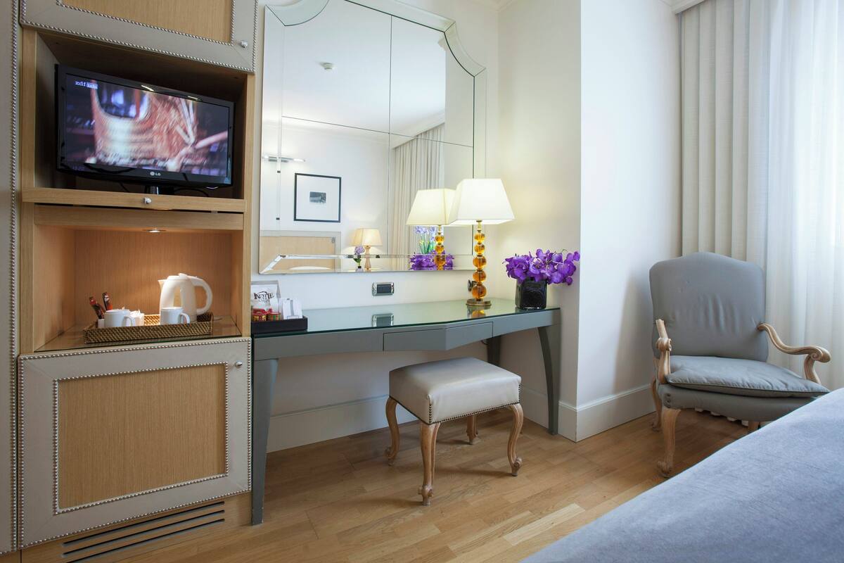 A hotel room with a television, a desk with a lamp, a chair, and a small table with a vase of flowers.