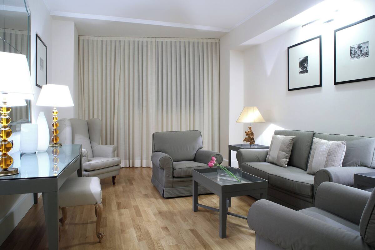 A modern living room with a grey sofa, a small table, and a chair. There is a window with vertical blinds, a lamp on the table, and framed pictures on the wall.