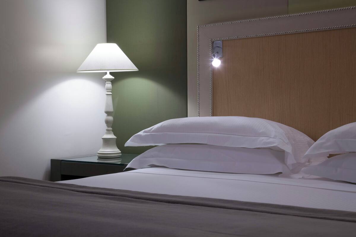 A neatly made bed with white linens and pillows, a white table lamp on a bedside table, and a wooden headboard with a decorative trim.