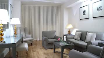 Ein modernes Wohnzimmer mit grauem Sofa, einem kleinen Tisch und einem Sessel. Ein Fenster mit vertikalen Jalousien, eine Lampe auf dem Tisch und gerahmte Bilder an der Wand sind zu sehen.