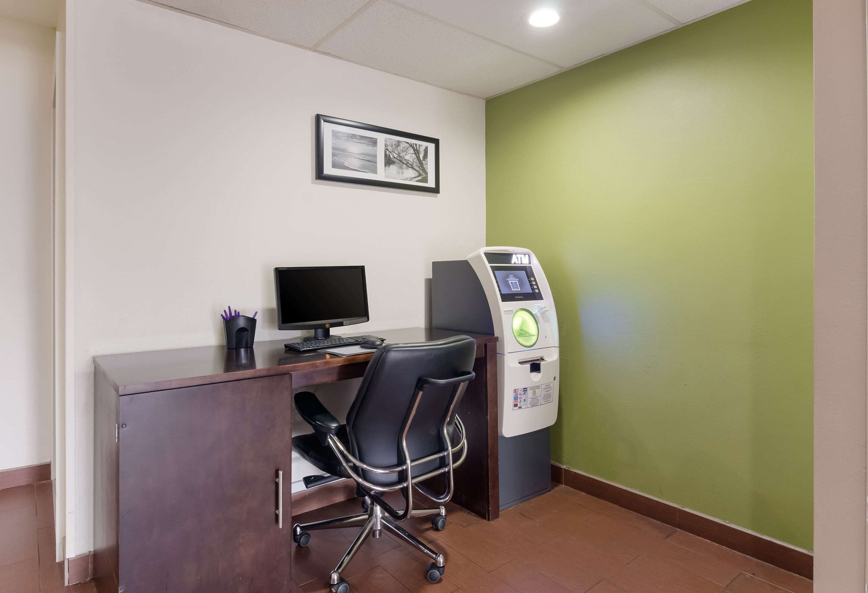 A room with a desk, chair, computer, ATM, and a framed picture on the wall.
