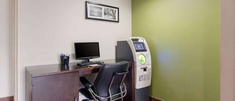 A room with a desk, chair, computer, ATM, and a framed picture on the wall.