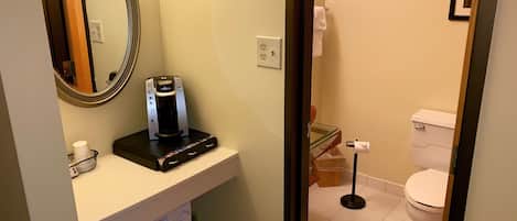 A bathroom with a coffee maker, a mirror, a toilet, and a trash bin.