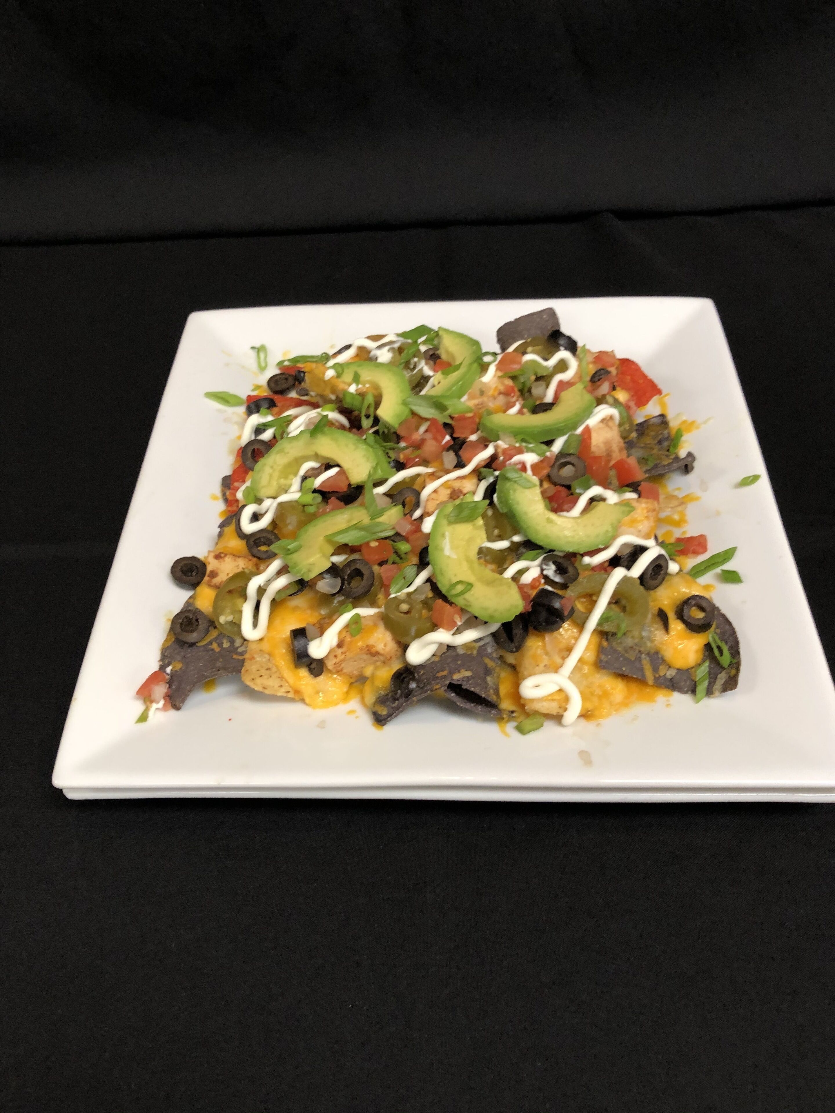 Um prato quadrado branco com nachos cobertos de queijo, azeitonas pretas, pimentões verdes e um molho branco.
