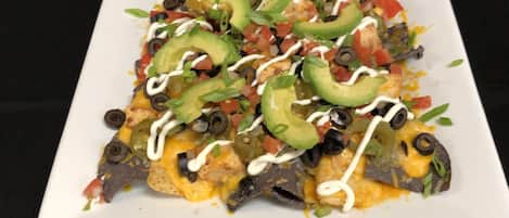 A square white plate with nachos topped with cheese, black olives, green peppers, and a white sauce.