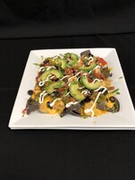 Un plato cuadrado blanco con nachos cubiertos de queso, aceitunas negras, pimientos verdes y salsa blanca.