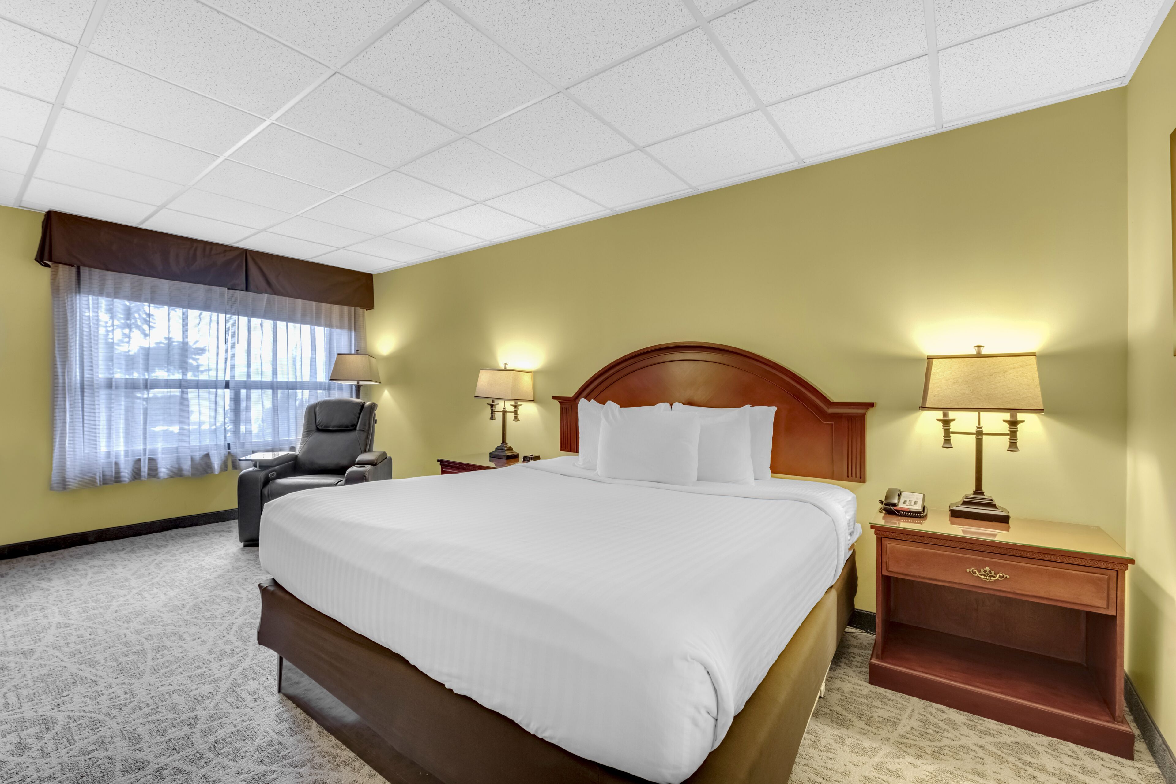 Quarto de hotel com uma cama grande, duas mesas de cabeceira com luminárias, uma cadeira e uma janela com cortinas leves.