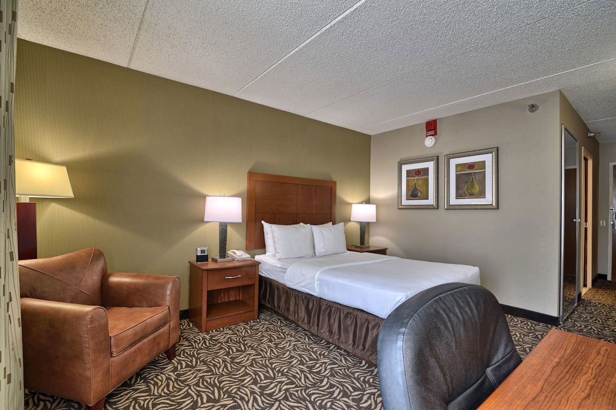 A hotel room with a bed, two bedside lamps, a telephone, a brown leather chair, a wooden nightstand, and a patterned carpet.