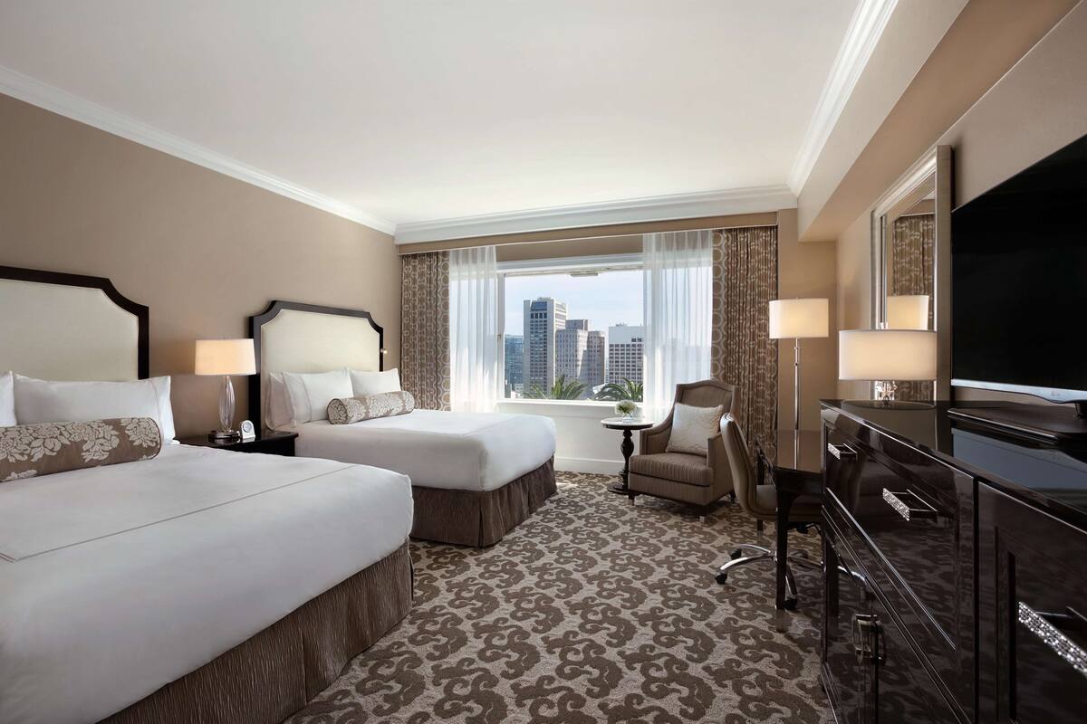 A hotel room with two beds, a TV, a desk, and a view of the city skyline.