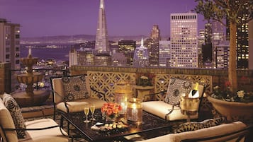 A rooftop terrace with a city skyline view, seating area with cushions, a table with drinks, and decorative elements.