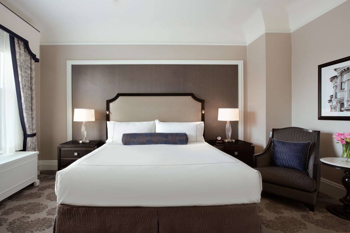 A hotel room with a large bed, two bedside tables with lamps, a chair, and a small table with a vase of flowers.