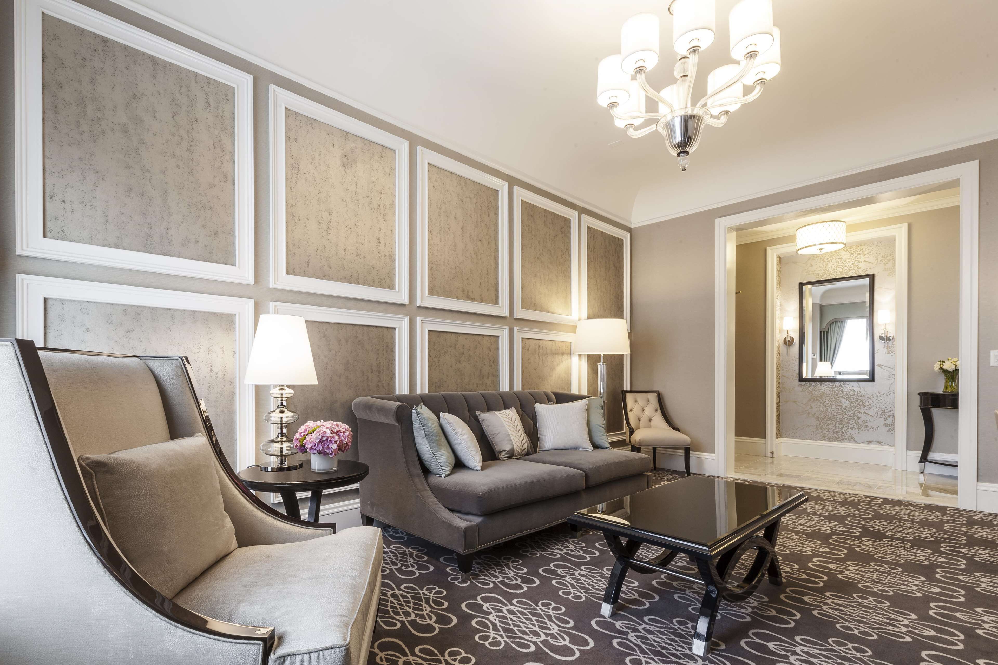 A modern living room with a grey sofa, a black coffee table, and a beige armchair. There is a chandelier above the sofa and a lamp on a side table.