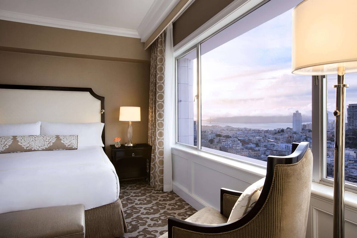 A hotel room with a large bed, a chair, a nightstand with a lamp, and a view of a cityscape through a large window.