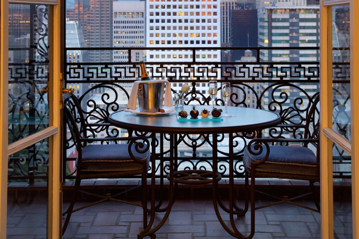 A balcony with wrought iron railings, a round table with chairs, and a view of a city skyline with tall buildings.