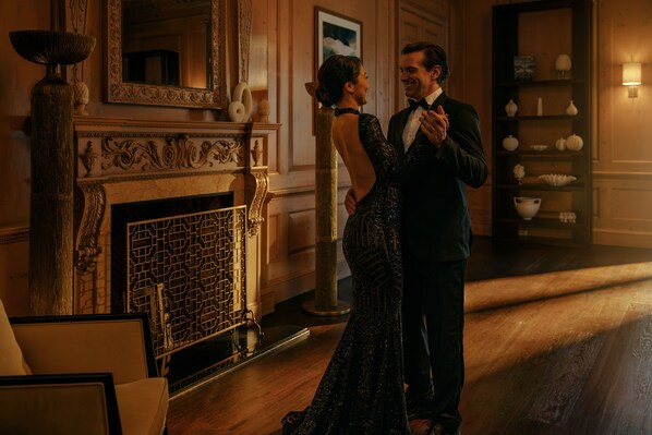 Una pareja baila en una habitación cálidamente iluminada con chimenea, espejo ornamentado y una repisa decorativa.