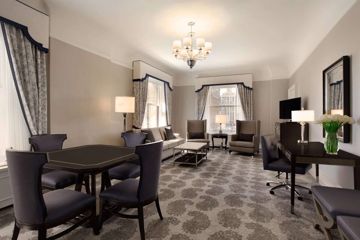 A hotel room with a dining table, chairs, a sofa, a TV, and a chandelier.