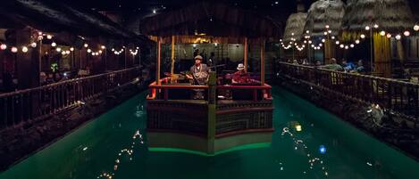 A boat-shaped bar with a thatched roof, surrounded by a water canal with string lights.