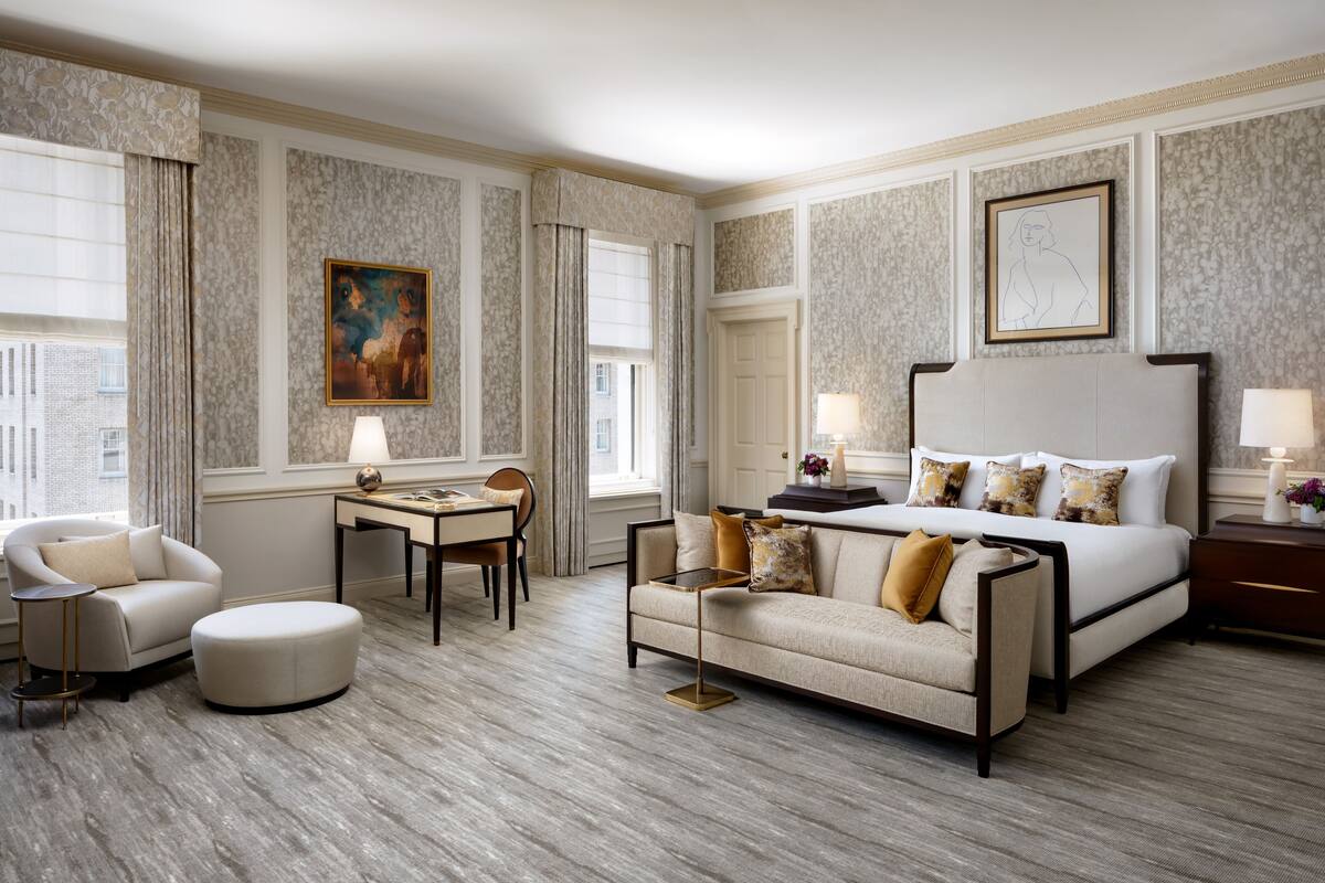 A hotel room with a large bed, a sofa, a desk, and a chair. There are two windows with curtains, a painting on the wall, and a lamp on the desk.