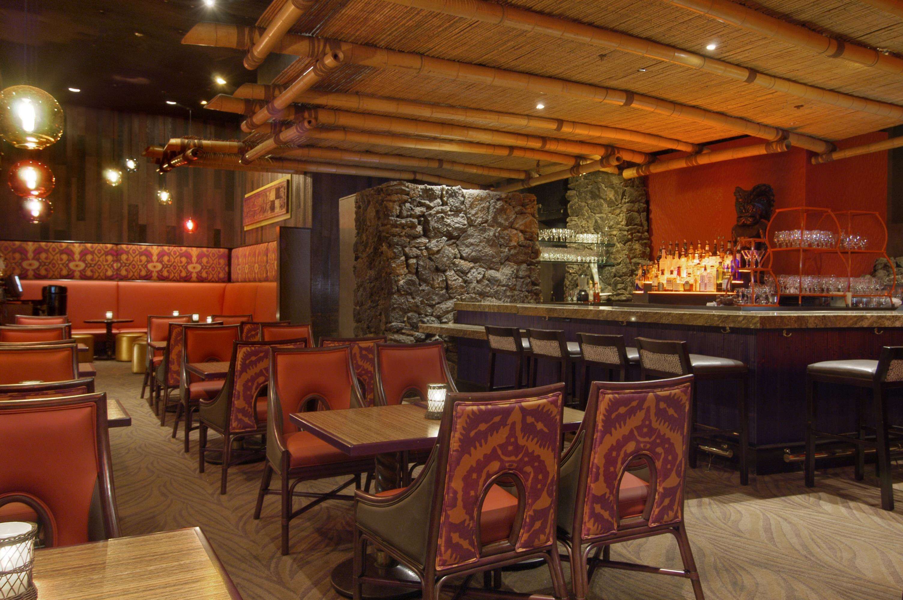 Ein Restaurant mit Sitzbänken und Tischen, ein Barbereich mit Steinwand sowie eine Auswahl an Flaschen und Gläsern auf Regalen.