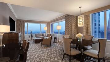 A hotel room with a large window, a dining table with chairs, a sofa, and a TV.
