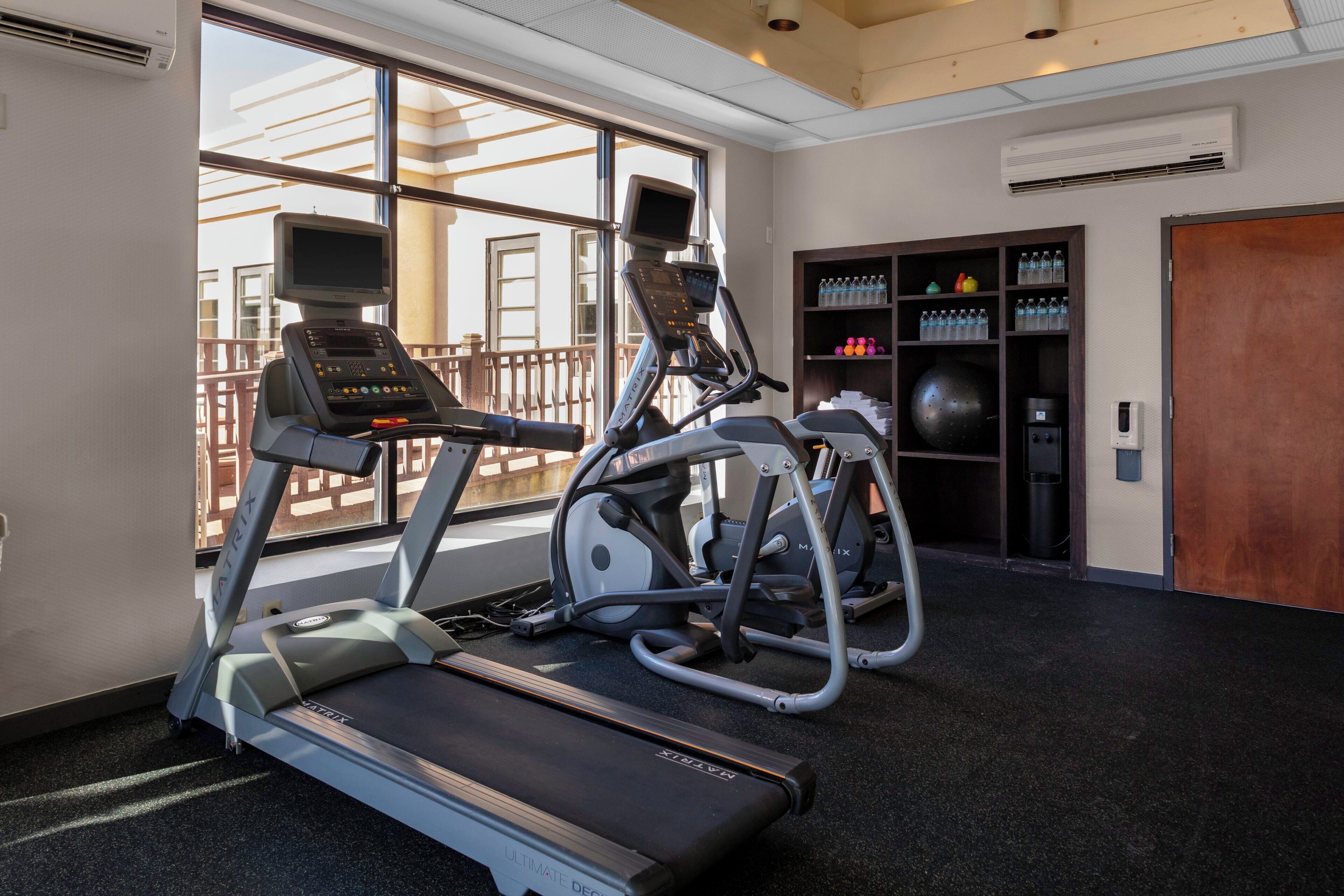 A gym with treadmills, an elliptical, and a stationary bike, with a view of the outdoors through large windows.