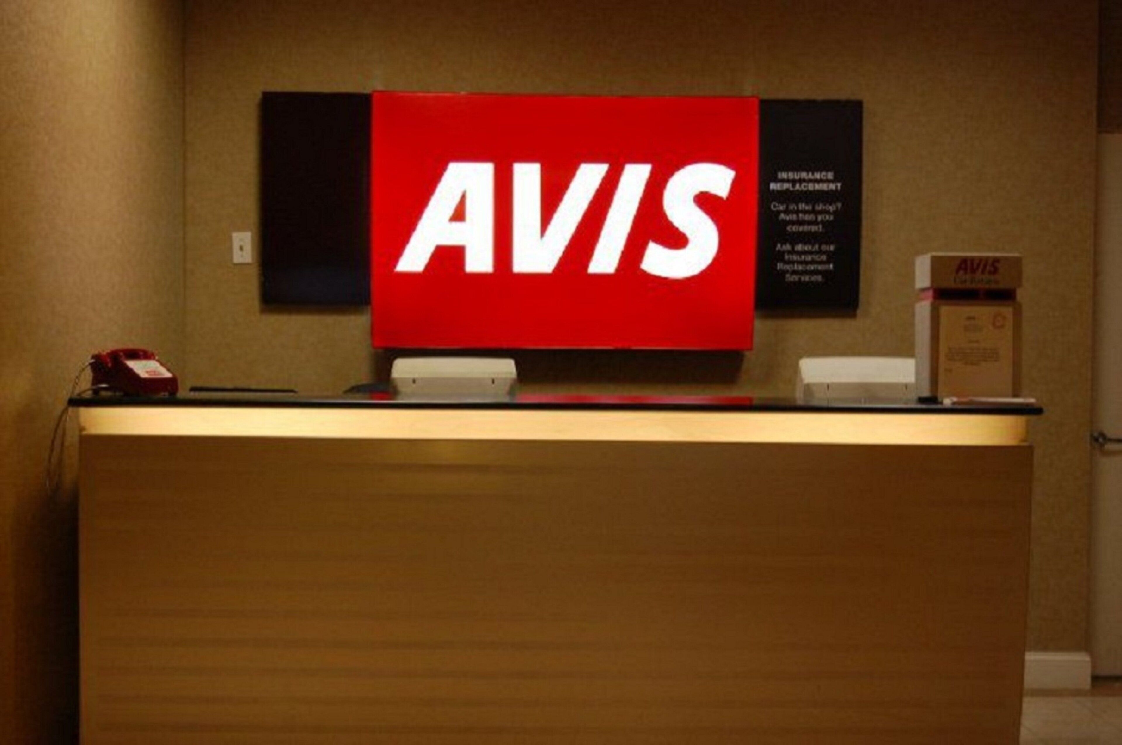 Un mostrador de recepción con un gran cartel de AVIS, un teléfono y una pequeña caja con la marca AVIS.