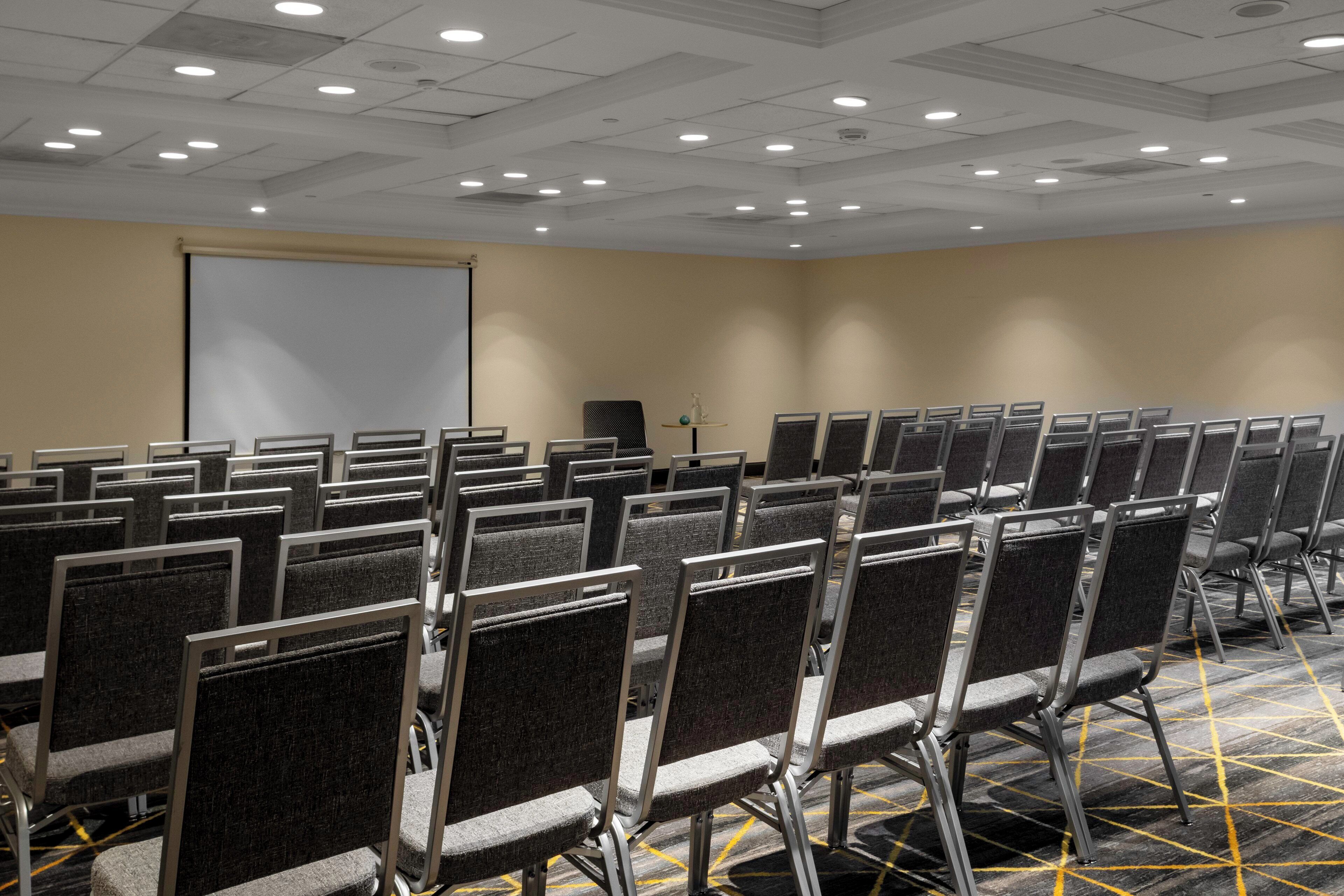 A conference room with rows of chairs, a projector screen, and a carpeted floor.
