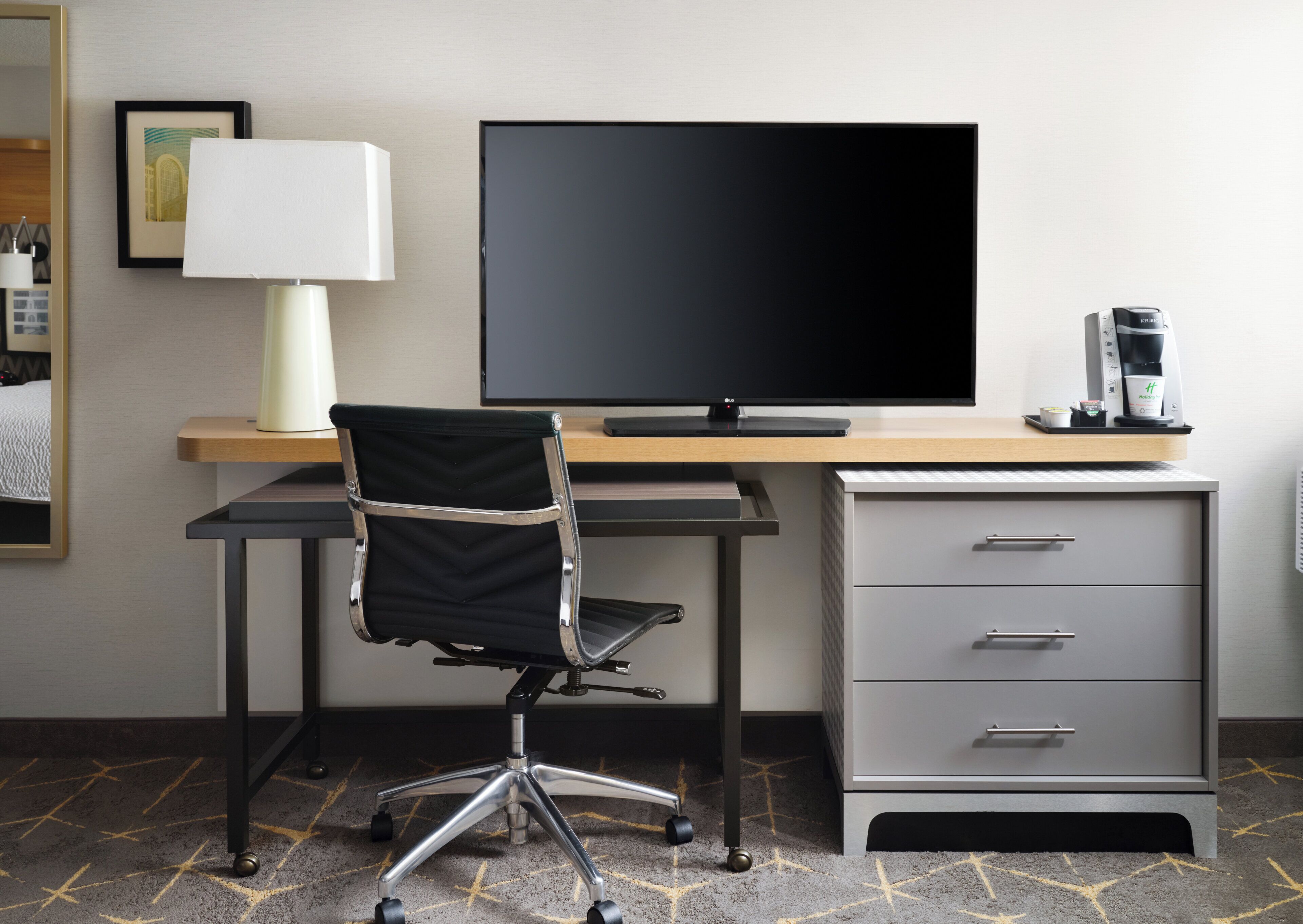 A hotel room with a desk, a chair, a flat-screen TV, a lamp, a coffee maker, and a filing cabinet.