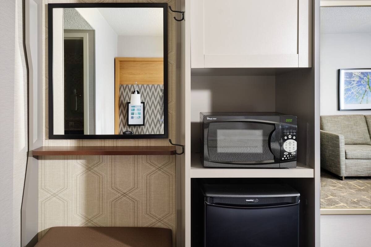 A compact kitchenette with a microwave, a small refrigerator, and a mirror above a wooden shelf.