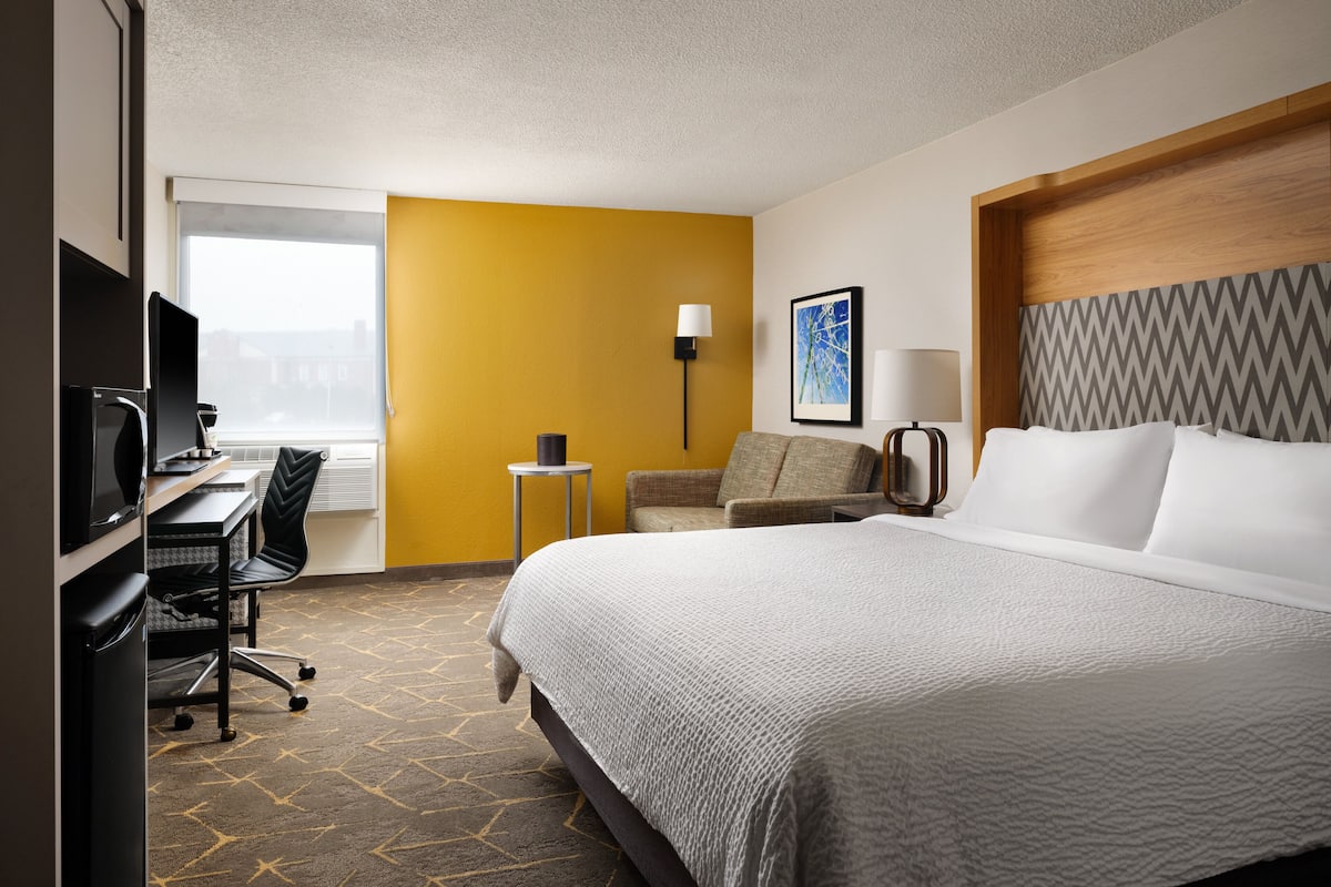 A hotel room with a bed, a desk with a computer, a chair, a lamp, and a window.