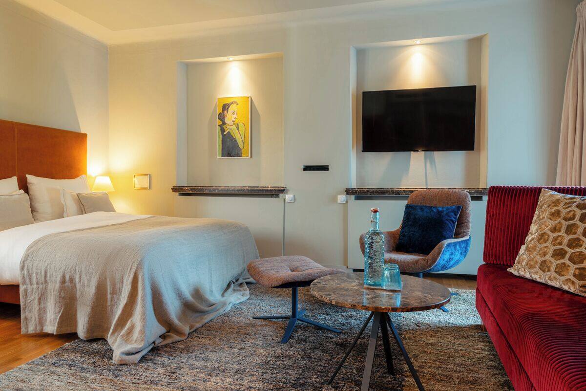 Una habitación de hotel con una cama, un sofá rojo, un televisor montado en la pared y un cuadro.
