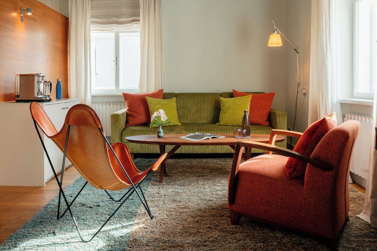 Una sala de estar acogedora con un sofá verde, sillones rojos, una mesa de centro y una mesita auxiliar. Hay un radiador debajo del sofá y una ventana con cortinas.