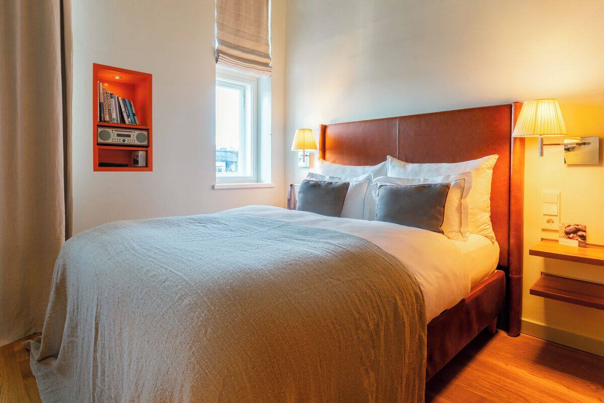 Una habitación de hotel con una cama grande, dos lámparas de noche, una estantería y un ventanal.