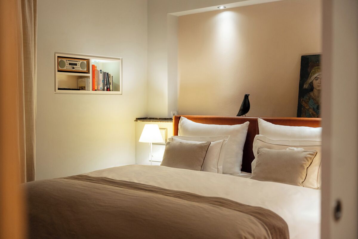 Ein Hotelzimmer mit einem ordentlich bezogenem Bett, einem Bücherregal mit Büchern, einer Lampe und einem Bild an der Wand.