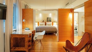 A modern hotel room with a wooden desk, a comfortable chair, a bed with a large window, and a painting on the wall.