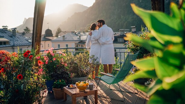 A couple embracing on a balcony with potted plants, a table with fruit, and a view of buildings and mountains in the background.