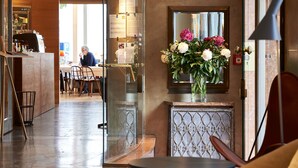 A hotel lobby with a reception desk, a mirror, a vase with flowers, and a view of the dining area.