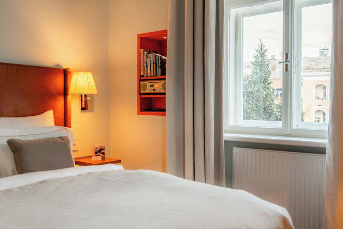 Habitación de hotel con cama, lámpara de noche, estantería y ventana que ofrece vistas a edificios y árboles.