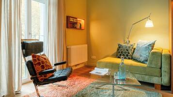 Un salon chaleureux avec un fauteuil en cuir, un canapé vert orné de coussins à motifs, une table basse en verre et un lampadaire.
