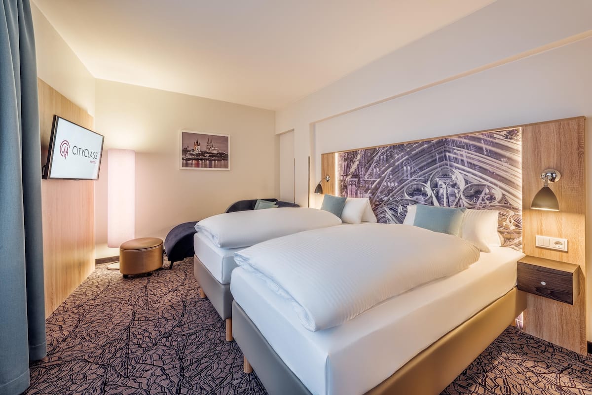 Habitación de hotel con dos camas, un escritorio, una silla, un televisor y una obra de arte con un paisaje urbano en la pared.