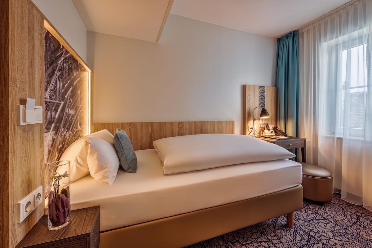 Habitación de hotel con cama, mesita de noche, lámpara y vistas al exterior.