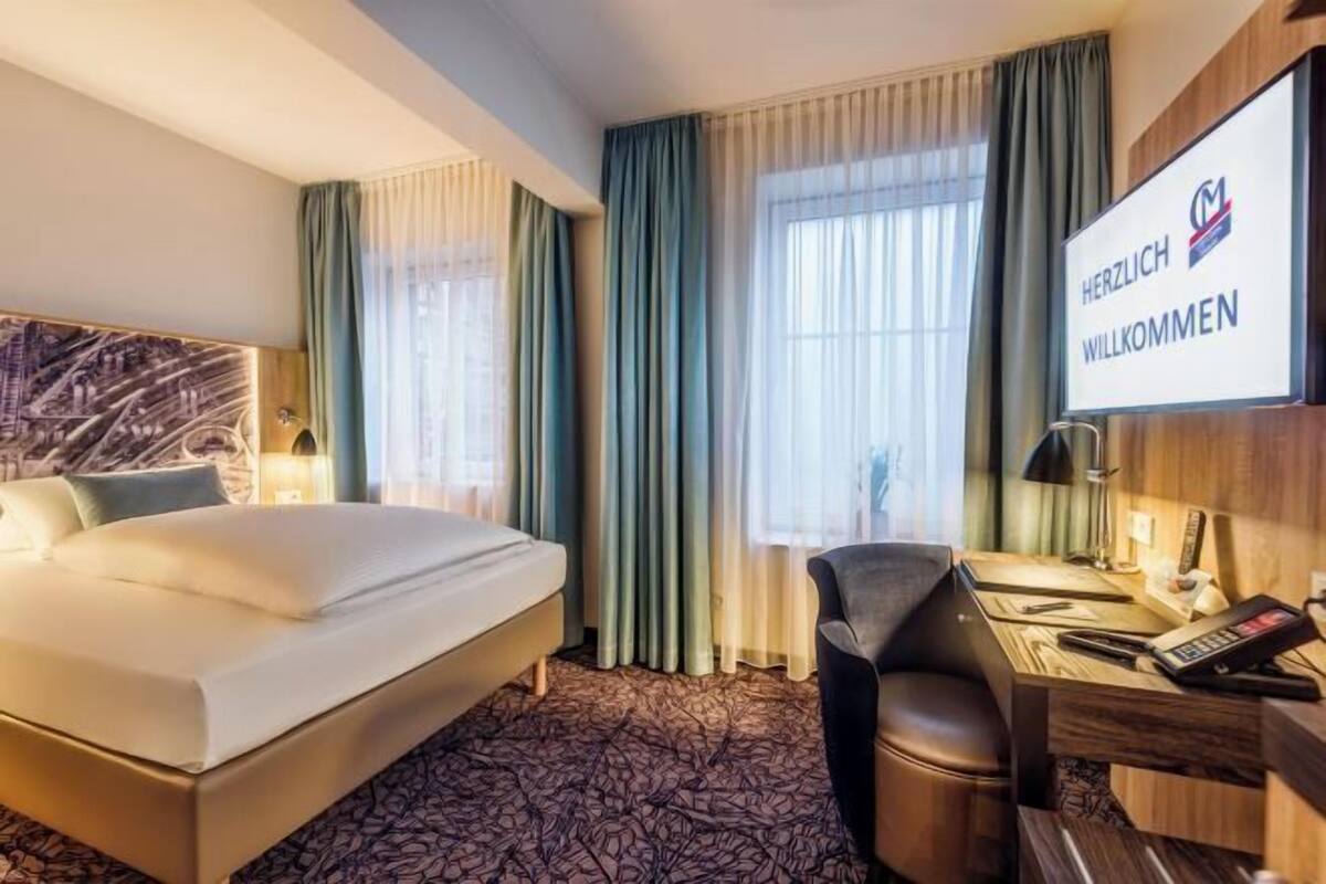 Habitación de hotel con cama, escritorio, silla, televisor y cortinas.