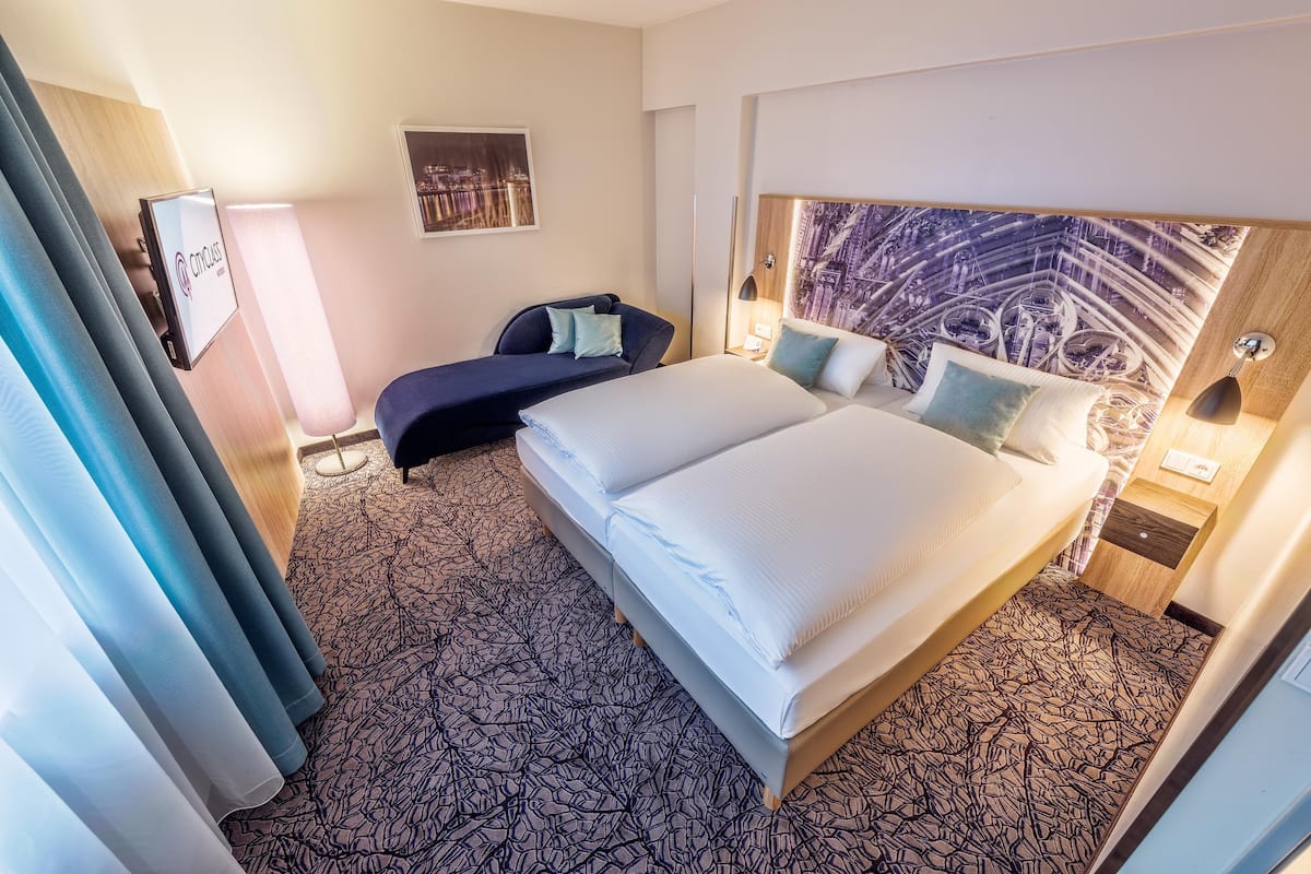 Habitación de hotel con una cama grande, un escritorio y una silla. Cuenta con ventana con cortinas, televisor montado en la pared y cabecera con diseño de paisaje urbano.