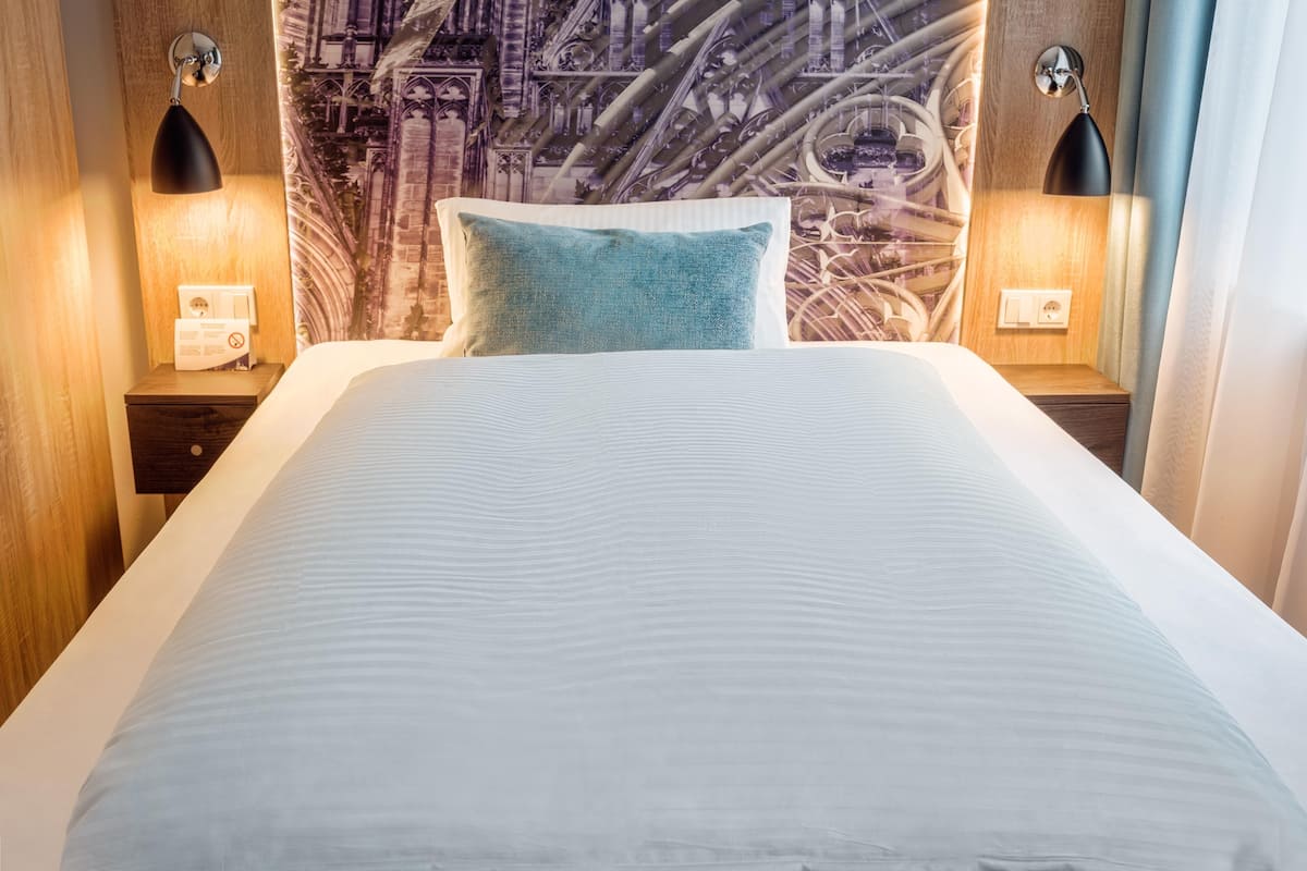 Una cama bien hecha con un almohá de blanco y azul, dos mesitas de noche con lámparas, y una pared con un mural detallado.